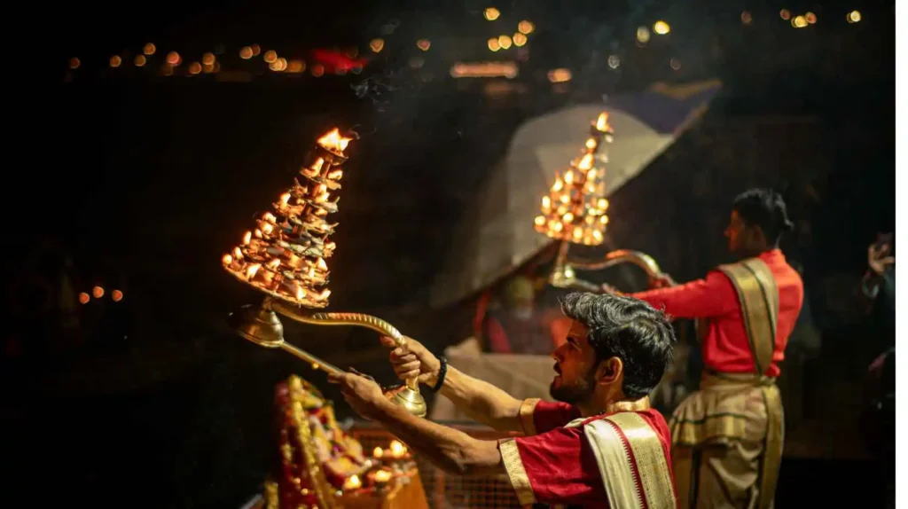 Priests in red and gold attire raising tiered brass oil lamps during the evening Ganga Aarti on a Varanasi ghat