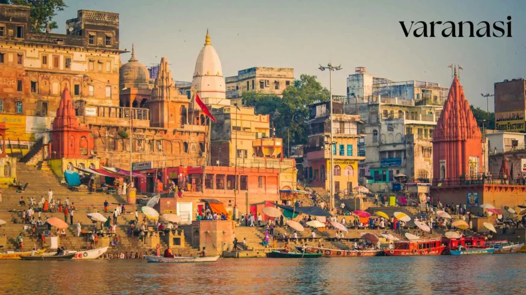 Pilgrims and shaded stalls on the stone steps of a Varanasi ghat, framed by red sandstone and white-domed Hindu temples